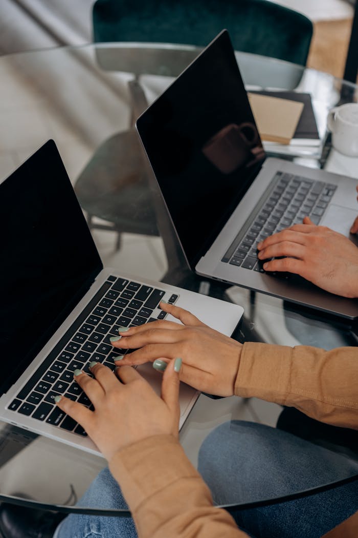 Two people working on laptops at a glass table in an indoor setting.